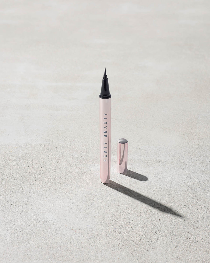 The Flyliner Longwear Liquid Eyeliner standing upright on a light grey concrete surface. The eyeliner is open, revealing its bold black tip, while its exterior features a sleek light metallic pink finish.